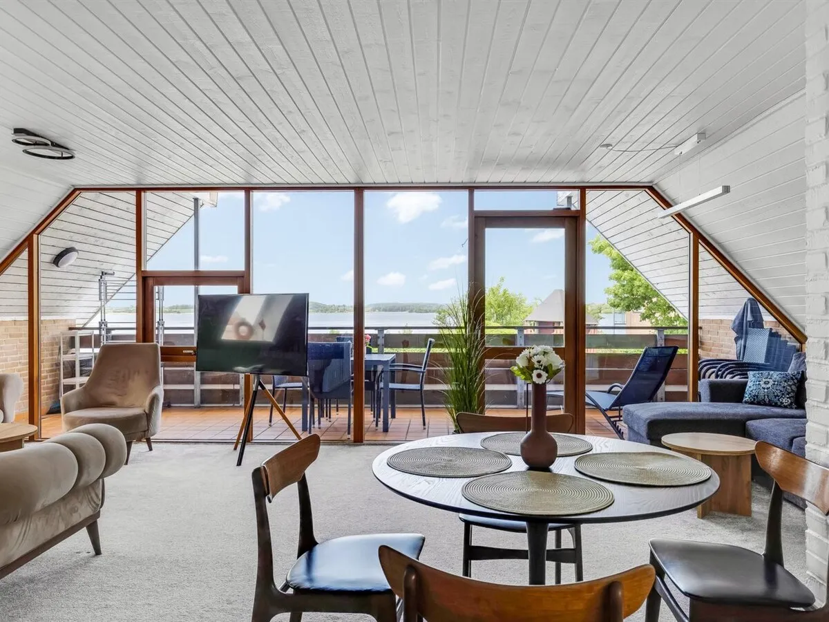 Stuen med spisebord, tv og panoramavinduer mod balkonen og Hejlsminde Nor